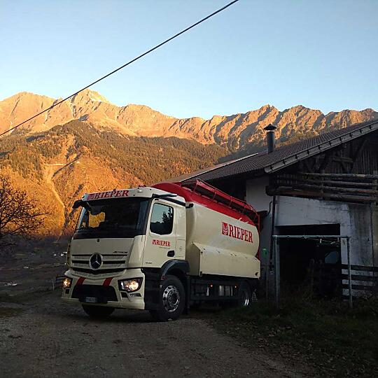 Ein Rieper LKW bei einem Bauernhof und im Hintergrund Berge, die in der Abendsonne leuchten.