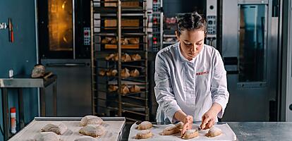 A baker is placing several loaves of bread on a baking tray in a bakery.